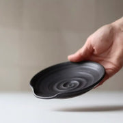 Hand holding a Gravesco Pottery handmade spoon rest, featuring a matte-black finish with a spiral pattern and a slight pouring spout, ideal for kitchen use.