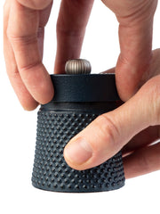 Hands twist the top of the Peugeot | Bali Cast Iron Pepper Mill, showcasing its textured design and metal adjustment knob, filled with aromatic Sichuan pepper.