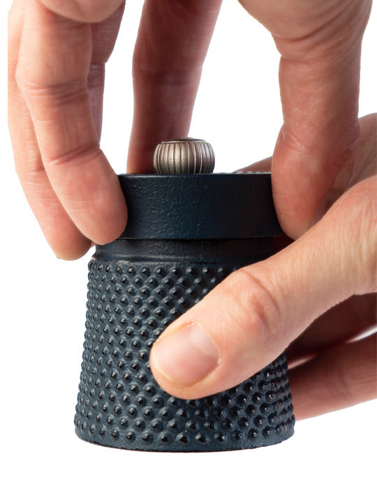 Hands twist the top of the Peugeot | Bali Cast Iron Pepper Mill, showcasing its textured design and metal adjustment knob, filled with aromatic Sichuan pepper.