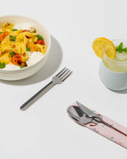 W&P - Stainless Steel Utensils Set in Silicone Carry Case next to a bowl of pasta and drink, highlighting compact, durable utensils for travel or work.