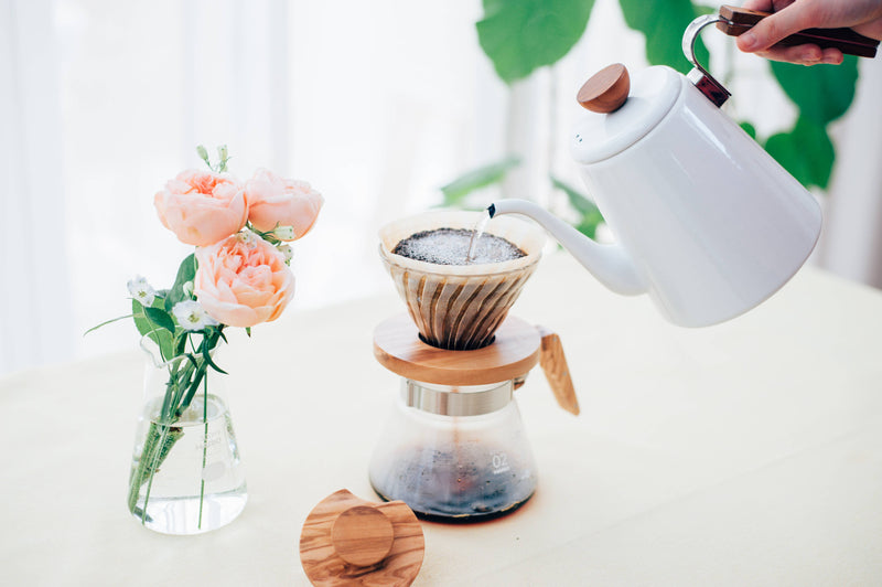 HARIO USA - HARIO V60 Glass Dripper 02: Olive Wood, showcased with a pour-over coffee setup, highlighting its conical shape and wooden collar in a bright, airy setting.