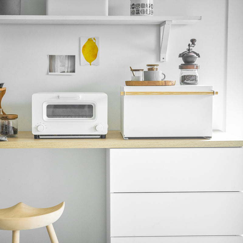 Yamazaki Home's Simple Steel Bread Box in White + Wood on a minimalist kitchen counter with a toaster-oven, coffee grinder, and decorative tray.