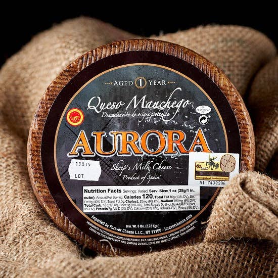 Nicola's Marketplace - DOP Aurora Manchego Kosher Cheese Wedge: 6 ounces, a whole wheel with branded rind, sits on burlap, showcasing its semi-firm texture.