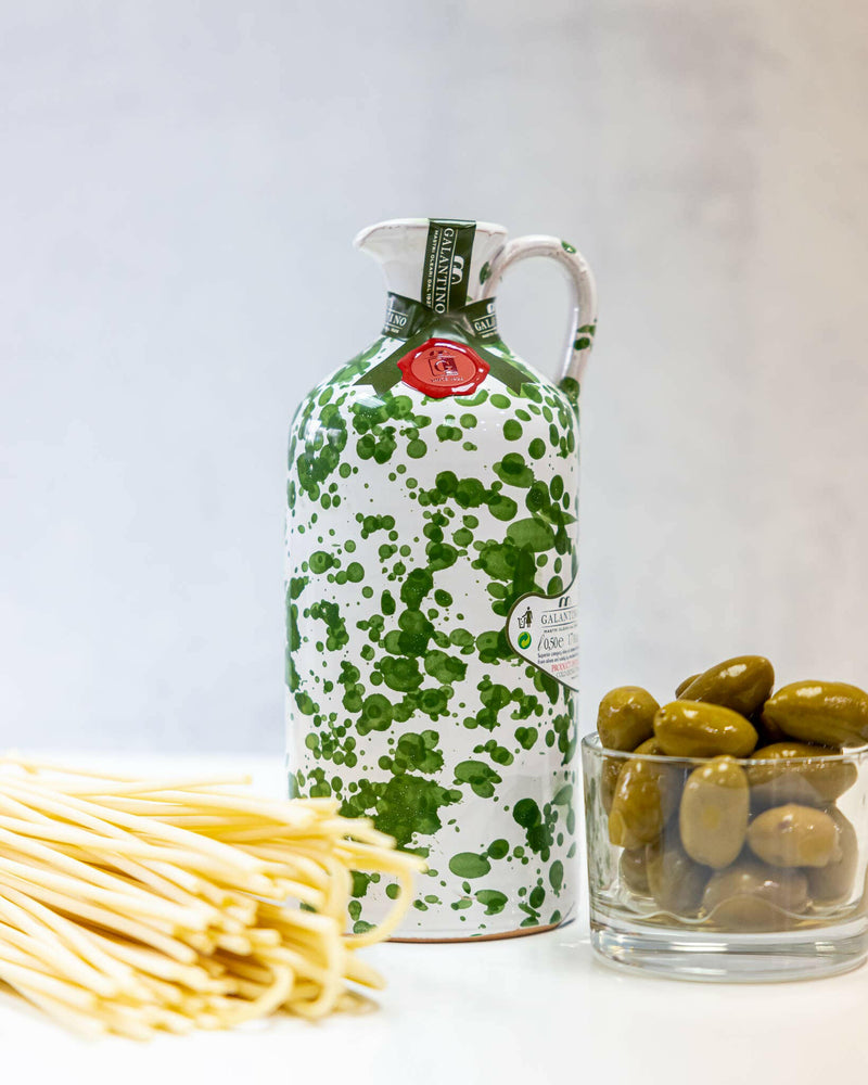 Frantoio Galantino Extra Virgin Olive Oil in a hand-painted ceramic jug, featuring green spots, alongside a glass of green olives and a red wax seal.