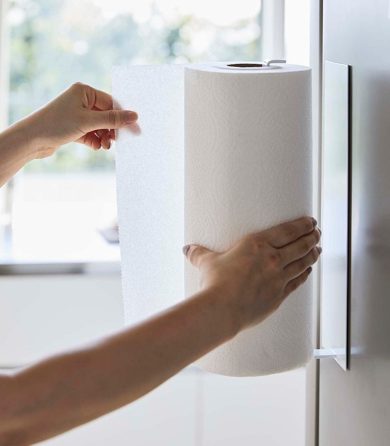 Yamazaki Home's Magnetic Steel Paper Towel Holder in use, mounted on a wall, as hands pull a sheet, showcasing its practicality and sleek design.