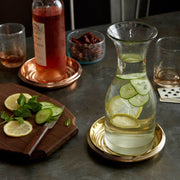 Sir|Madam Brass Bottle Coaster cradles a glass carafe on a brass tray, keeping the table dry amid a refreshing beverage setup with sliced lemons and cucumbers.