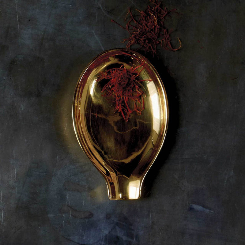 Sir|Madam Brass Spoon Rest on slate surface, showcasing its gold-plated, spoon-shaped design, cradling fine red saffron threads, embodying elegance and functionality for kitchen use.