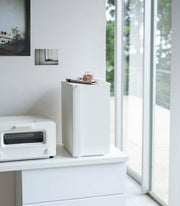 Yamazaki Home Bread Box, Vertical, Steel, Black sits elegantly in a minimalist kitchen nook, enhancing decor by reducing countertop clutter with its sleek design.