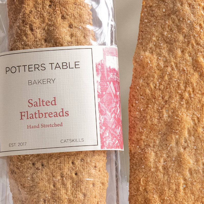 Potters Table - Salted Olive Oil Flatbreads, a packaged bakery item showing hand-stretched flatbreads; one in clear wrapper, one lying beside it, ideal for snacking.