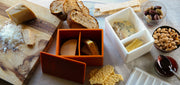 CapaBunga® Cheese Vault on a table with various cheeses, bread, crackers, olives, and preserves, showcasing versatile cheese storage for freshness and flavor preservation.