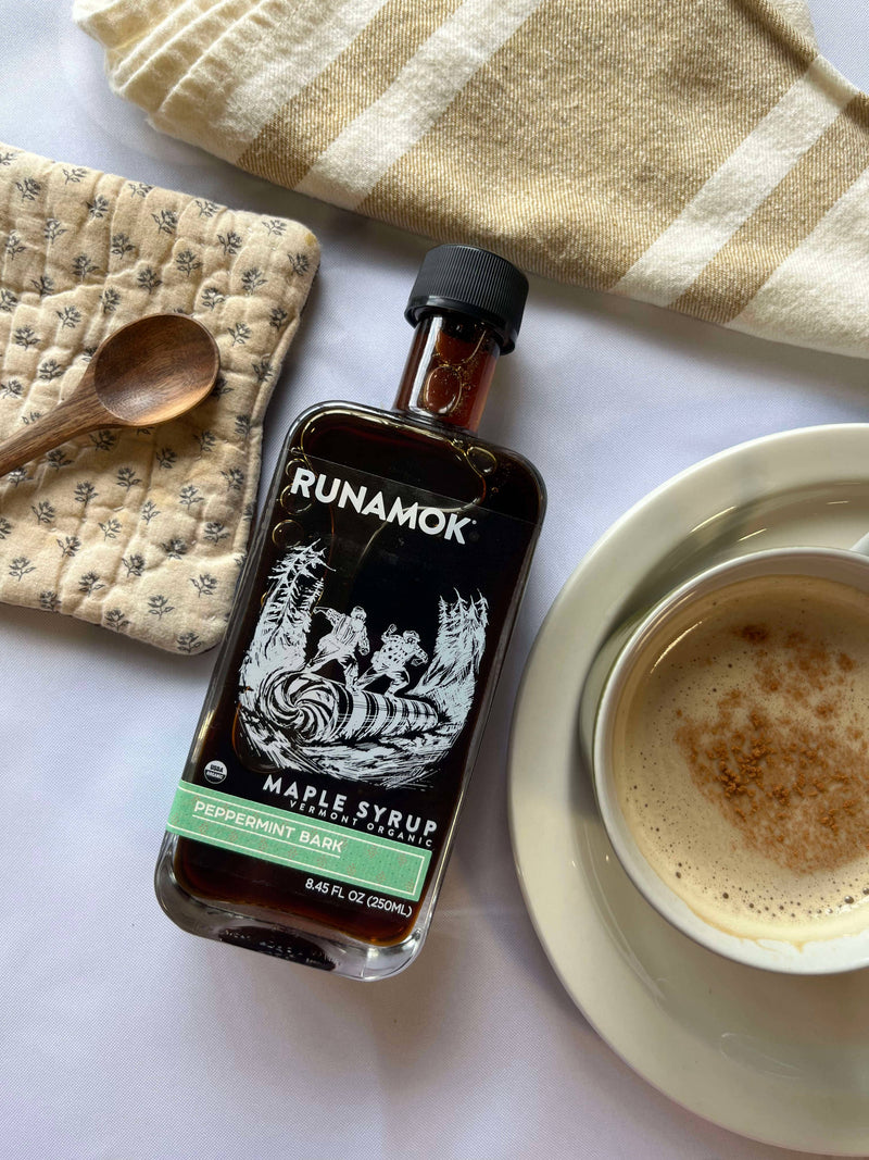 Runamok - Peppermint Bark Organic Maple Syrup 250ml, surrounded by a pot holder, wooden spoon, striped towel, and a frothy coffee cup on a saucer.