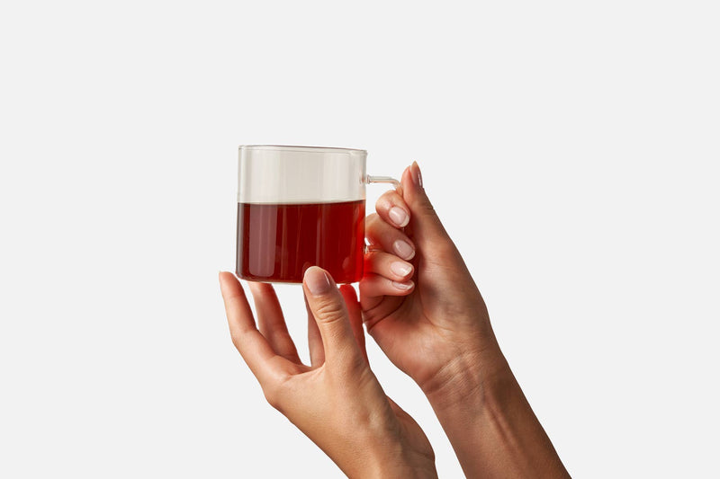 Hands holding a clear glass mug with a dark amber liquid, showcasing Big Heart Tea Co. - Rosy & Bright 10 ct Tea Bags, featuring hibiscus, rose, and cinnamon blend.