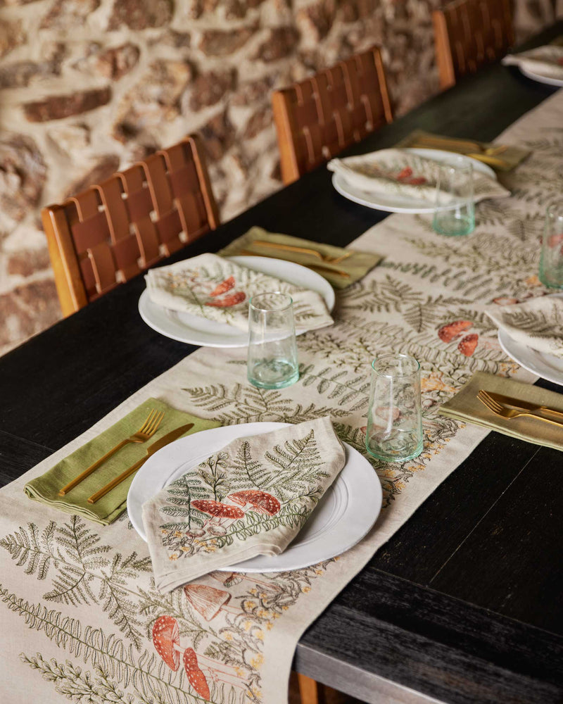 Coral & Tusk - Mushrooms and Ferns Bouquet Dinner Napkin folded on plates with matching table runner, gold flatware, and green-tinted glasses on a rustic table.