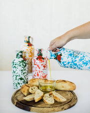 Hand pours olive oil from a white and blue Zia Pia - Fantasia Extra Virgin Olive Oil Ceramic-Yellow by Galantino cruet onto bread, showcasing artisanal craftsmanship.