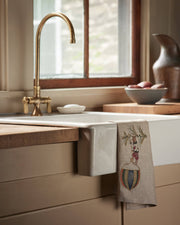 Coral & Tusk - Deck the Halls Bear Tea Towel draped over a white apron-front sink, featuring whimsical embroidery in a light-filled kitchen setting.