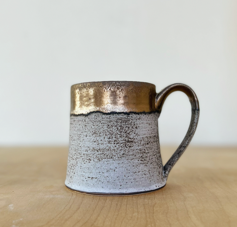 Jessica Wertz Ceramics - Grey + Gold Mug, featuring a speckled ceramic body and metallic bronze rim and handle, sits on a wooden surface. Holds 12-14 ounces.