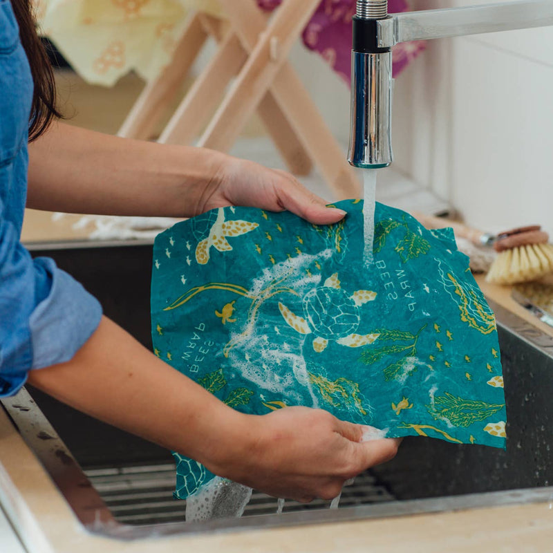 Bee's Wrap - Assorted 3 Pack - Oceans: A person washing a turtle-patterned reusable fabric wrap in a sink, illustrating its washable, sustainable design.