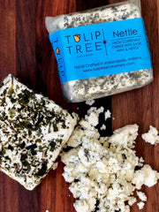 Tulip Tree Creamery - Nettle cheese wedge on a wooden board, featuring herb-infused slices and crumbles. Perfect picnic pairing with sauvignon blanc.
