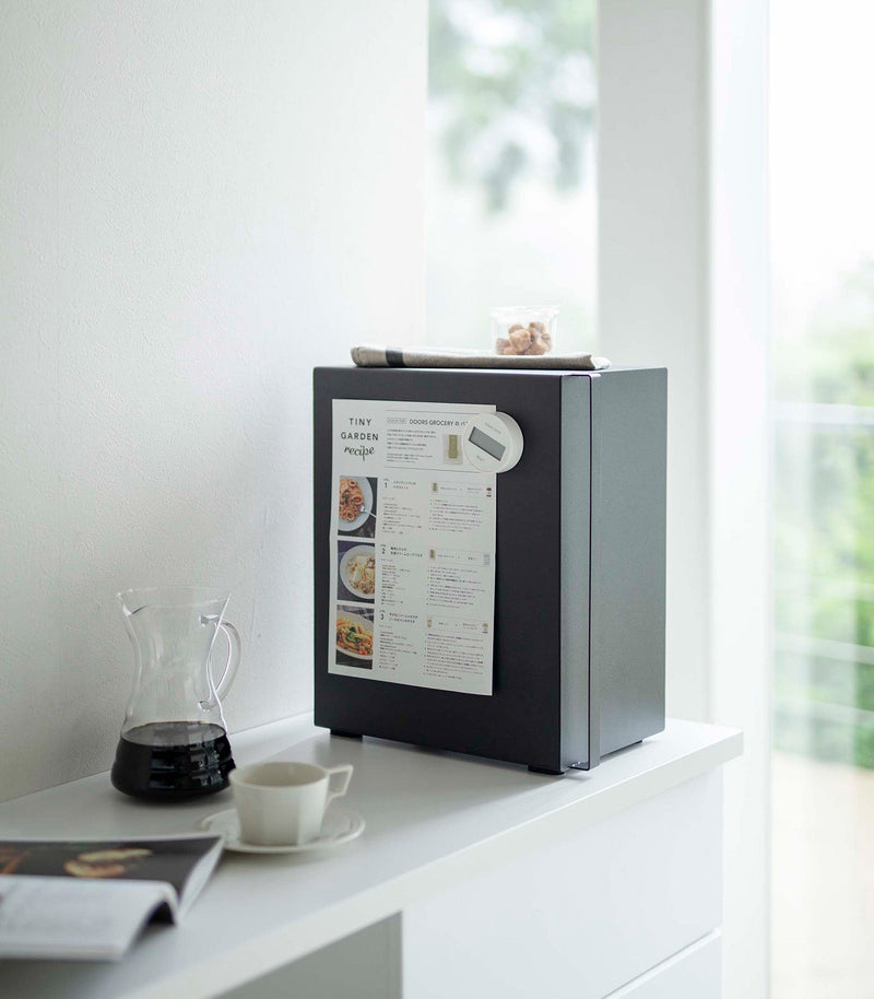 Yamazaki Home | Bread Box in matte-black on a white sideboard, featuring a handle and hinged side. Holds a recipe magnet, with snacks and coffee nearby.