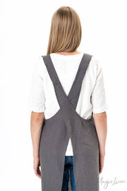 Woman in a grey apron, wearing a white shirt, styled as MagicLinen’s pinafore cross-back linen apron, featuring a twin-pocket design, ideal for cooking and gardening.