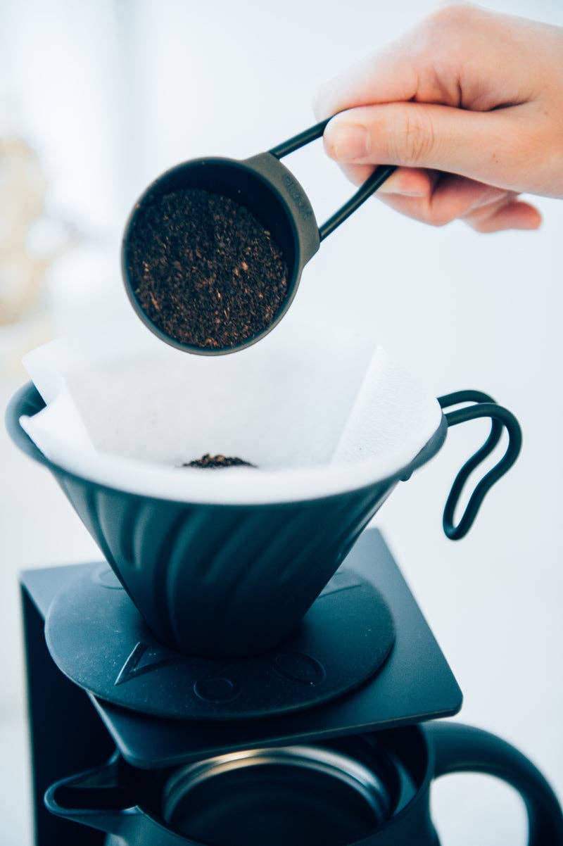 Pour-over coffee setup with HARIO USA - V60 Metal Dripper: Silver. A hand holds ground coffee over a spiral-ribbed dripper atop a carafe.
