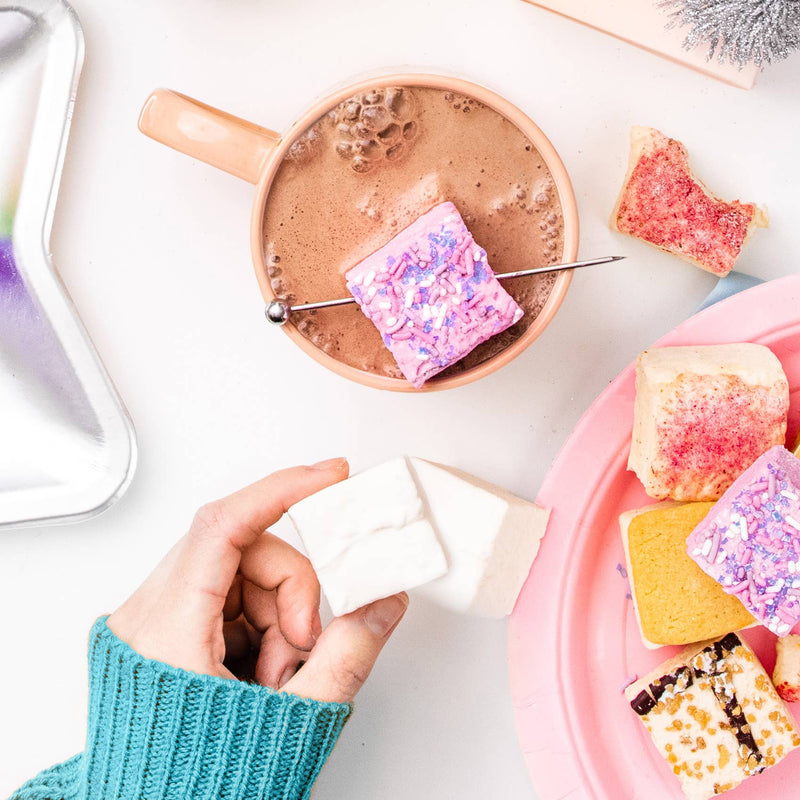 Cozy hot-cocoa scene featuring Malvi Mallow Cotton Candy 4-Pack Marshmallows, artistically sprinkled and skewered, surrounded by a delightful marshmallow variety on a pink plate.