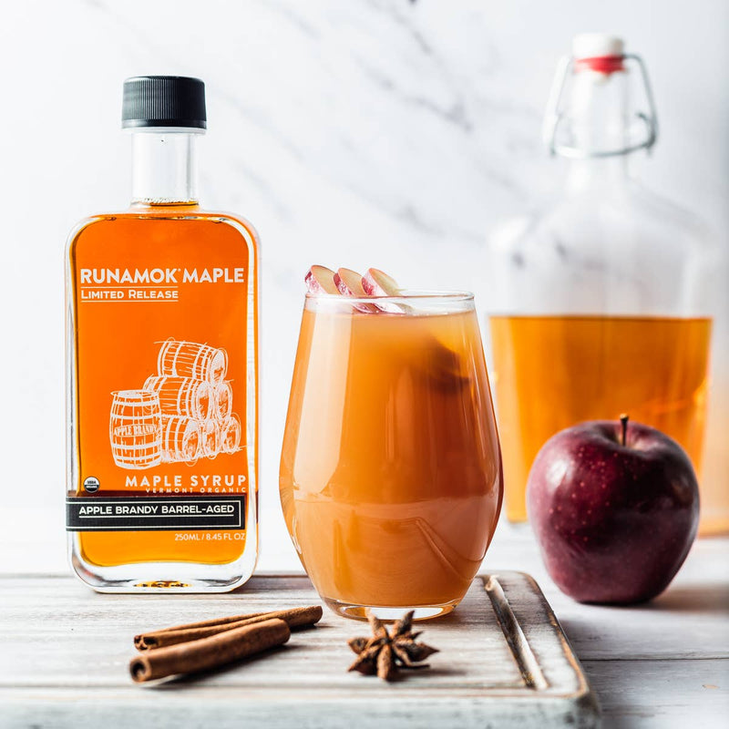 Runamok - LIMITED RELEASE Apple Brandy Barrel-Aged Organic Maple Syrup with cocktail setup, featuring apple slices, cinnamon, star anise, and apple, suggesting a cozy autumn drink.