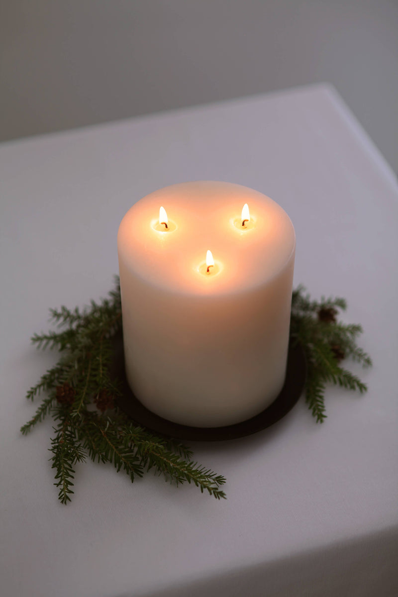 Mo&Co Home - 200h Burn | GRAND Beeswax Pillar Candle: A large, ivory pillar candle with three lit wicks, adorned with evergreen sprigs on a dark tray.