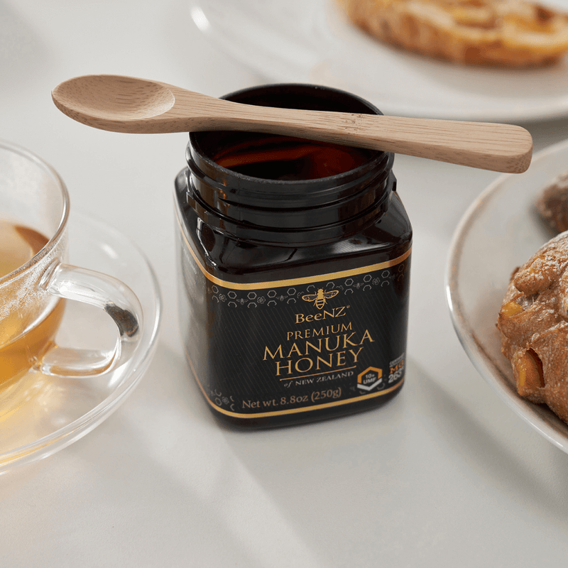 BeeNZ Premium Manuka Honey with a bambu® Bamboo Tea Spoon atop, flanked by tea and pastries, ideal for eco-friendly kitchen and gift setups.