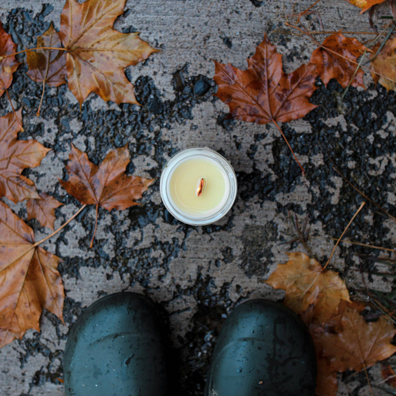 Among the Flowers - Essential oil candle | HOLIDAY: Hearth, unlit, on damp pavement with scattered maple leaves, next to dark green rubber boots.