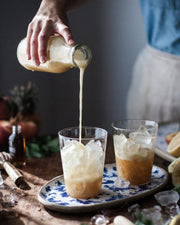 Pouring frothy drink into plastic cup set on floral platter with Farmhouse Pottery Glasses nearby; showcases borosilicate glassware's luxury and practicality for Athens Cooks customers.