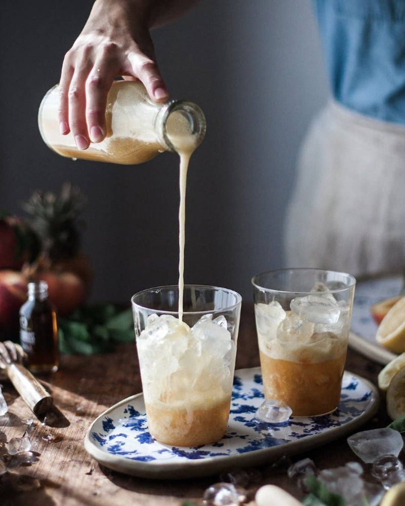 Pouring frothy drink into plastic cup set on floral platter with Farmhouse Pottery Glasses nearby; showcases borosilicate glassware's luxury and practicality for Athens Cooks customers.
