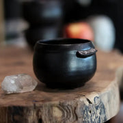 Gravesco Pottery - Black Cauldron Bowl: A handcrafted, matte black ceramic bowl, reminiscent of witchcraft, alongside a clear quartz crystal on a wooden slab.