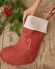 MagicLinen | Candy Cane Embroidered Christmas Stocking, held by two hands, features a natural cuff and candy-cane motif, laying on rustic wood with festive greenery accents.