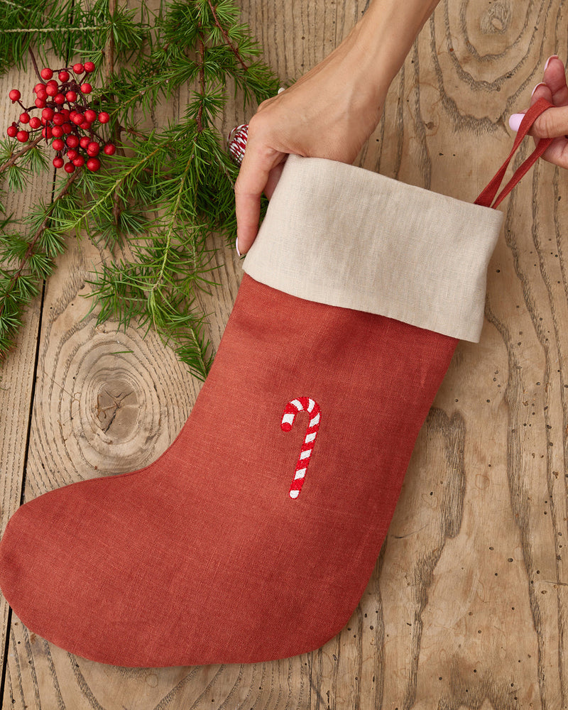 MagicLinen | Candy Cane Embroidered Christmas Stocking, held by two hands, features a natural cuff and candy-cane motif, laying on rustic wood with festive greenery accents.