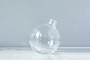 Etú HOME | Clear Sphere Bud Vase, a transparent, artisanal glass vase designed for single-stem displays, elegantly sits slightly off-center on a white surface.