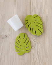 Graf Lantz Merino Wool Monstera Coaster Solid 4 Pack displayed with a white cup and green felt leaves on a wooden surface.