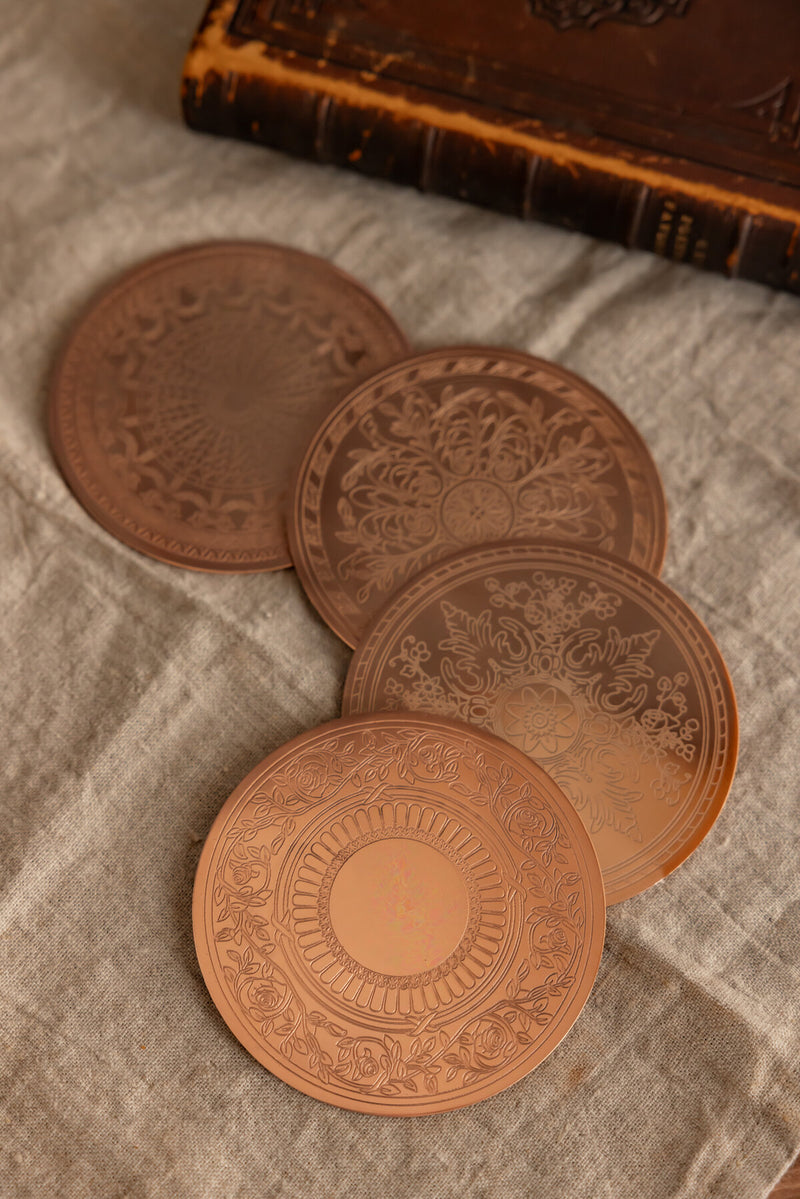 Coppermill Kitchen LLC | Vintage Inspired Coasters - Set of Four, embossed copper discs with intricate patterns, displayed on linen, reflecting European elegance and collectible charm.