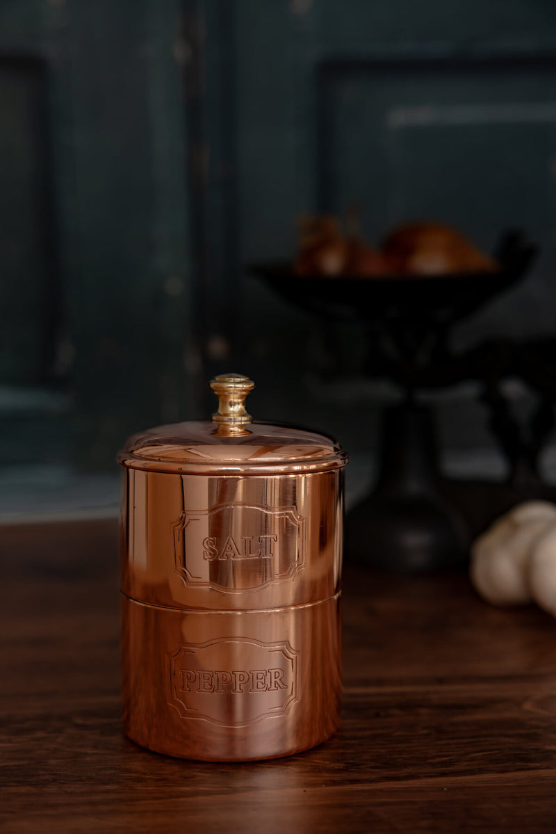 Coppermill Kitchen LLC | Vintage Inspired Salt & Pepper Cellar set, two-tiered copper canisters with brass knobs, reflecting European kitchen charm on a wooden surface.