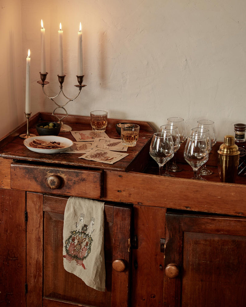 Rustic sideboard with drinks setup, featuring Coral & Tusk | Togetherness Tea Towel embroidered with rabbits and wreath, alongside candelabra, glasses, and cocktail accessories.