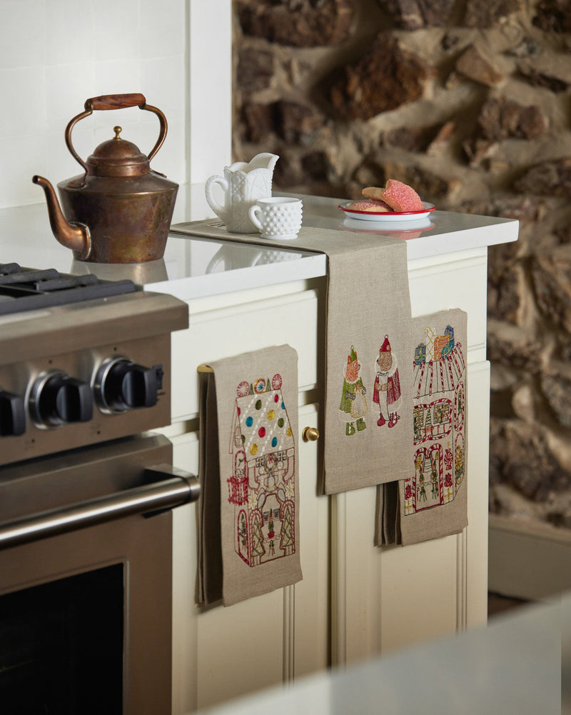 Coral & Tusk | Cookie Workshop Tea Towel embroidered with festive motifs drapes over a cabinet, complementing a copper kettle and holiday treats on a kitchen counter.