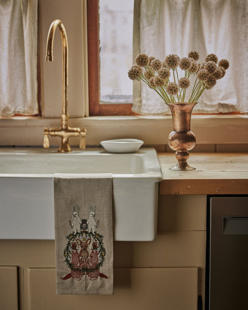Coral & Tusk | Togetherness Tea Towel with embroidered woodland animals and foliage, draped over a rustic farmhouse sink in a cozy kitchen setting.