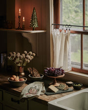 Togetherness Tea Towel with forest animal motif draped over a butcher-block counter, surrounded by a cozy kitchen display of cheeses, fruits, and snacks.