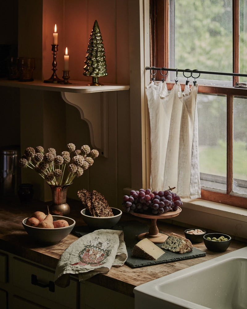 Togetherness Tea Towel with forest animal motif draped over a butcher-block counter, surrounded by a cozy kitchen display of cheeses, fruits, and snacks.