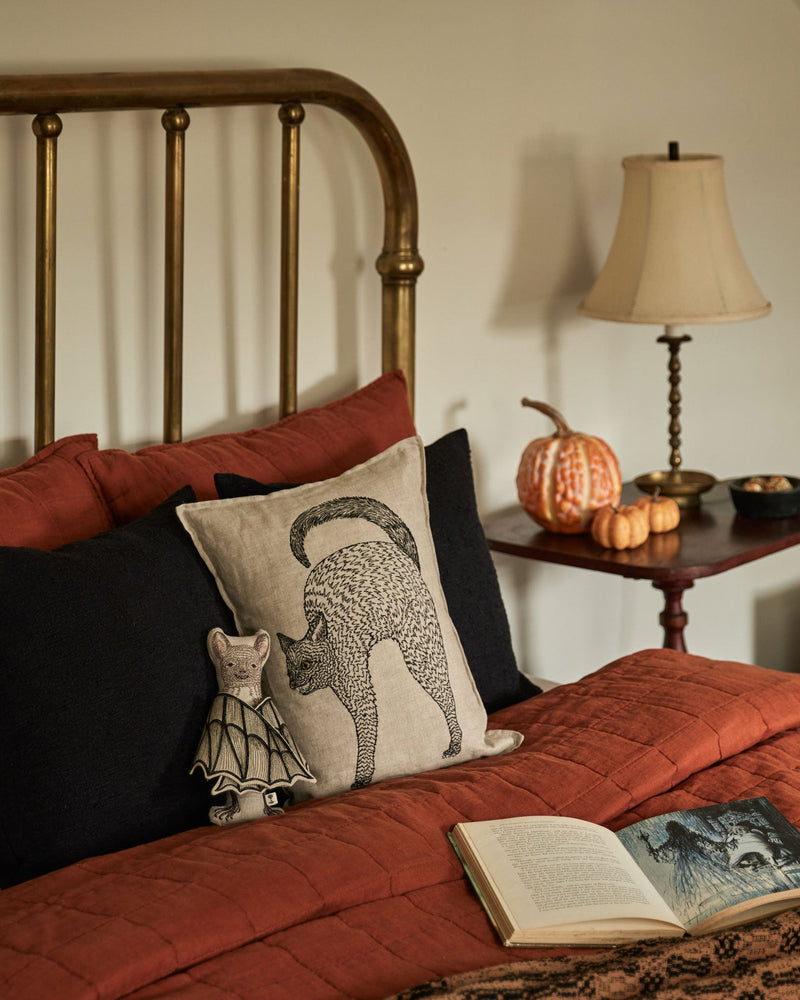 Coral & Tusk | Bat Surprise Doll nestled on a rust-orange quilted bed, with wings closed, amidst autumn-themed décor including pillows, a lamp, and decorative pumpkins.