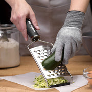 Person grating zucchini using a Microplane box grater while wearing Microplane Cut Resistant Kitchen Safety Gloves, ensuring safety and efficiency in culinary tasks.