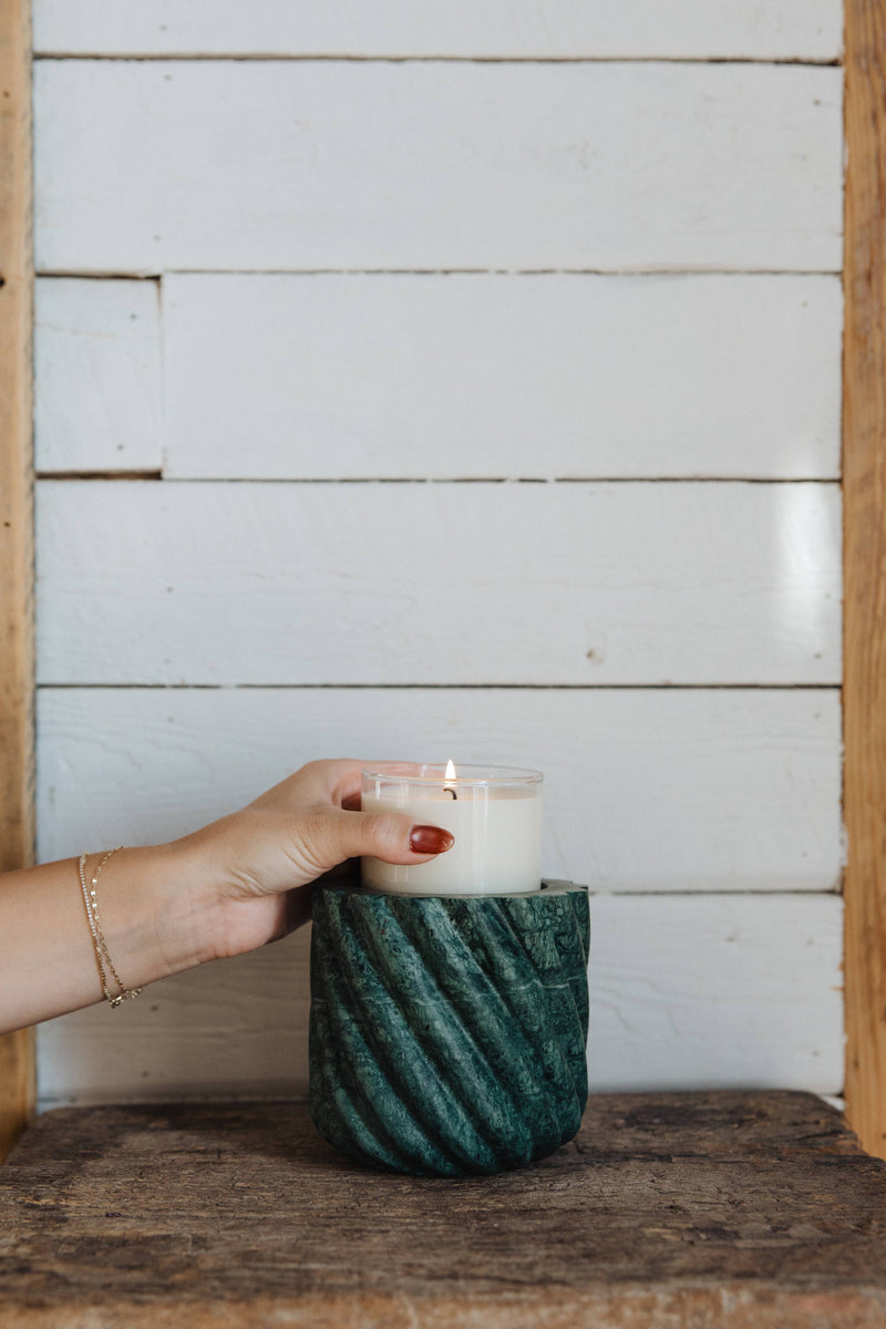 Hand with red nails holds lit Last Candle Co - Emerald Green Marble Refillable Candle in ribbed marble holder on wooden surface, showcasing its unique design.