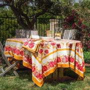 Couleur Nature - Pomegranate Yellow & Red tablecloth, 71 x 71, on a wooden table with matching napkins, pitcher, and glasses in a garden setting.