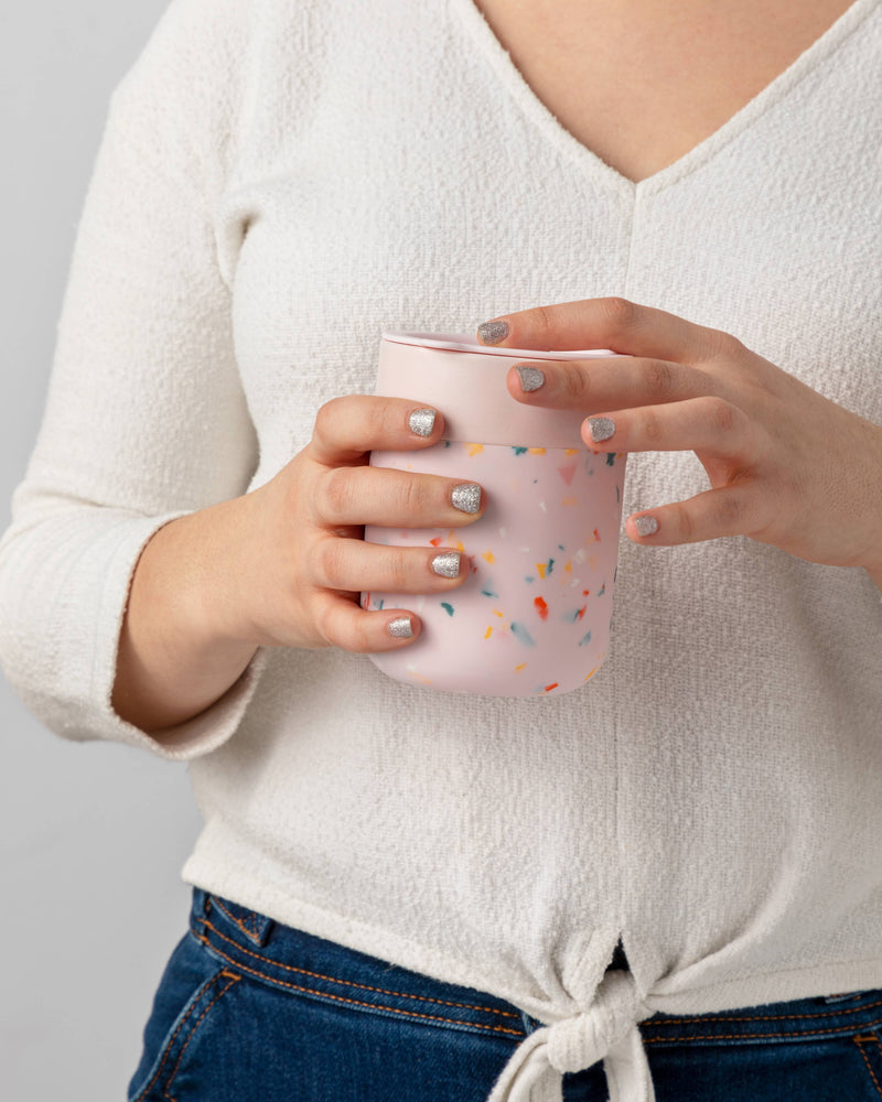 Person holding W&P - Reusable Ceramic Mug: Terrazzo Charcoal, featuring glittery nails. Mug has a splash-resistant lid and silicone sleeve for easy grip.