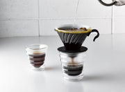 HARIO USA - V60 Metal Dripper: Silver in use, brewing coffee into a double-walled glass with hot water pouring from a kettle above.